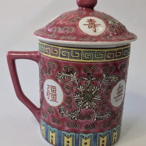 May include: A maroon ceramic mug with a lid, featuring intricate floral and geometric patterns. The mug has a handle and is adorned with Chinese characters in white circles. The lid has a small knob on top.