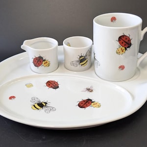 Può includere: Un set da colazione in ceramica bianca con una tazza, una lattiera, una tazzina e un vassoio ovale. Il set è decorato con coccinelle rosse e gialle, api e piccoli funghi rossi. Il vassoio ha una forma unica.