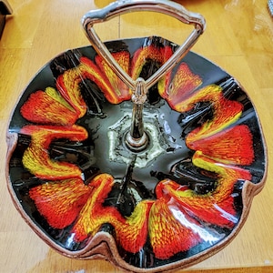 Puede incluir: Un plato de servir de cerámica negra con un asa plateada. El plato tiene un borde ondulado y festoneado y está decorado con un patrón de remolino de rojo, amarillo y naranja.