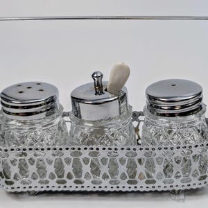 Puede incluir: Un soporte para condimentos de metal plateado con tres frascos de vidrio con tapas plateadas. Los frascos tienen un patrón de diamante y están llenos de especias. El soporte tiene un asa en la parte superior.