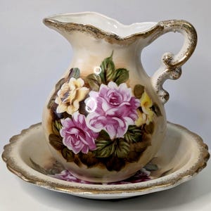 May include: A cream-coloured ceramic pitcher and basin set. The pitcher has a handle and is decorated with pink and yellow roses and green leaves. The rim and handle are trimmed with gold. The basin has a scalloped edge and a matching floral design.