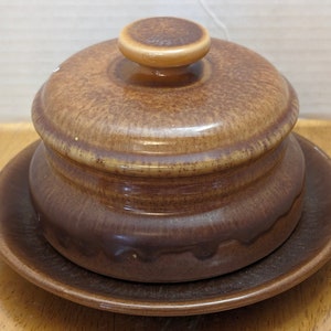 Peut inclure: Un beurrier en céramique brun avec un couvercle et une assiette assortie. Le beurrier a une surface texturée et une forme légèrement arrondie.