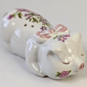 May include: White ceramic pig-shaped salt shaker with a pink bow and floral accents. The pig has closed eyes and a pink snout. Purple and green floral designs adorn the pig's body, adding a touch of whimsy to this kitchen accessory.