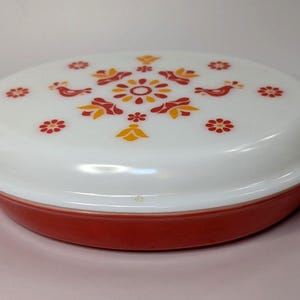 Könnte beinhalten: Ovale Auflaufform in Rot und Weiß mit Deckel. Der Deckel ist weiß und mit einem Blumen- und Vogelmuster in Rot und Gelb verziert. Die Form hat Griffe.