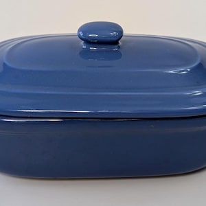 Op de afbeelding: Een blauwe keramische ovenschaal met een deksel. De schaal is ovaal van vorm met een handvat aan elke kant. Het deksel heeft een kleine, ronde knop. De schaal is ongeveer 30 cm lang.