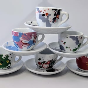 May include: A stack of six espresso cups and saucers with colorful abstract designs. The cups are white with splashes of color, including green, red, blue, and black.