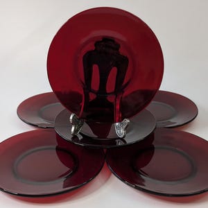 Puede incluir: Un juego de seis platos de vidrio rojo rubí. Los platos son redondos con un borde ligeramente elevado. Un plato se exhibe en un soporte decorativo plateado. Los platos están dispuestos sobre una superficie blanca.