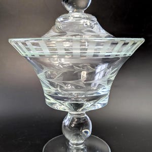 Könnte beinhalten: Transparente Bonbonniere aus Glas mit Deckel. Die Schale hat einen runden Sockel, einen bauchigen Stiel und eine ausgestellte Schüssel mit geätzten Blatt- und Streifenmustern. Der Deckel hat einen runden Knopf. Das Glas ist transparent.