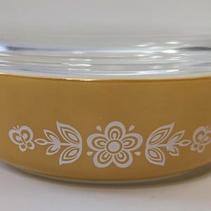 Könnte beinhalten: Eine Vintage Pyrex Auflaufform in Goldgelb, mit einem Deckel aus klarem Glas. Die Schüssel ist mit einem weißen Blumenmuster auf der Außenseite verziert. Fassungsvermögen ca. 1,4 Liter.