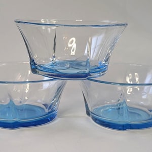 Puede incluir: Un conjunto de tres cuencos de cristal azul claro translúcido dispuestos en forma de pirámide. Cada cuenco presenta un borde ancho y acampanado, con una base interior lisa y curva y sutiles hendiduras verticales en los lados exteriores.