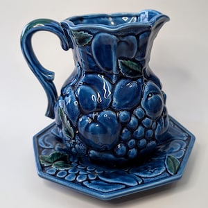 Peut inclure: Un pichet en céramique bleu et son dessous de plat hexagonal assorti. Le pichet est orné de fruits et de feuilles en relief. L'ensemble est bleu cobalt brillant, avec des accents verts sur les feuilles.