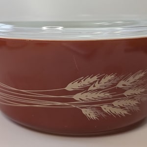 Könnte beinhalten: Eine braune Pyrex-Auflaufform mit einem Deckel aus klarem Glas. Die Schüssel hat ein Weizenmuster in Creme. Sie hat zwei Griffe und ist für den Ofen geeignet. Ein klassischer Küchenartikel.