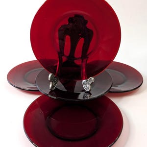 Può includere: Un set di piatti in vetro rosso rubino. Un piatto è esposto su un supporto decorativo argentato, con l'immagine di una sedia. Gli altri piatti sono impilati e disposti attorno al supporto. I piatti hanno un design semplice ed elegante.
