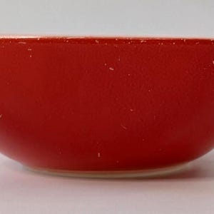 Puede incluir: Un pequeño cuenco de cerámica rojo con un acabado liso y brillante. El cuenco tiene forma redondeada y una base ligeramente blanquecina. El interior y el exterior son de color rojo vivo.