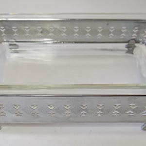 May include: A rectangular clear glass baking dish set inside a silver-toned metal frame. The metal frame features a repeating cutout pattern of small diamond shapes and has two side handles and four small feet at the base for stability.