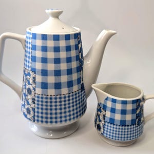 Puede incluir: Un juego de té de cerámica blanca que incluye una tetera y una jarra de crema. La tetera tiene tapa y un pico largo. Ambas piezas presentan un patrón de cuadros azules y blancos con detalles de patchwork floral. La jarra de crema tiene un asa pequeña y un diseño similar.