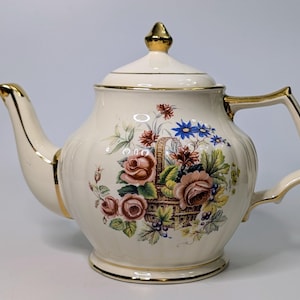 May include: A white teapot with a gold trim and floral design. The teapot has a round body with a curved spout and handle. The floral design features roses, blue flowers, and green leaves.