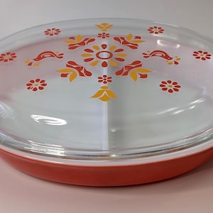 Könnte beinhalten: Ovale, geteilte Servierschale mit einem Deckel aus klarem Glas. Die Schale ist rot mit weißen Griffen. Der Deckel hat ein Blumendesign in Orange und Gelb. Ein Vintage-Küchenartikel.