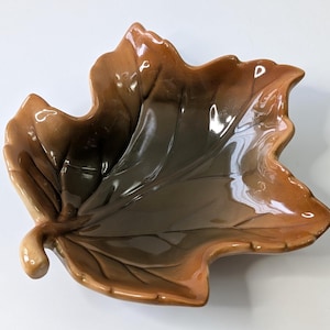 May include: A ceramic leaf-shaped dish with a glossy finish. The dish features a gradient of colors, transitioning from a light brown to a darker brown and green in the center. The dish has a detailed leaf vein pattern.