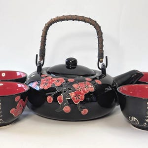 May include: A black ceramic tea set consisting of a teapot with a woven handle and four matching cups. The teapot features a red floral branch design. Each cup has a vibrant red interior and a black exterior decorated with white Chinese calligraphy.