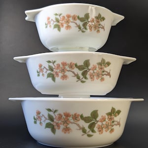 Op de afbeelding: Drie witte keramische kommen gestapeld, elk met een bloemmotief van perzikbloesems en groene bladeren op bruine takken. De kommen hebben aan weerszijden handvatten. De achtergrond is zwart.