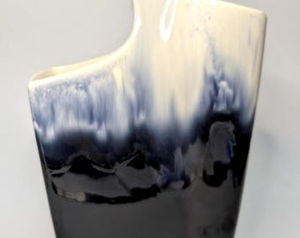 May include: A tall, ceramic vase with a unique shape and a striking color gradient. The top portion is a creamy white, transitioning to a deep navy blue and black at the base. The vase has a handle-like extension.
