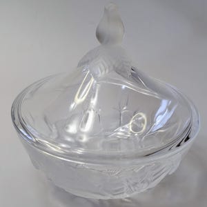 Puede incluir: Un cuenco de vidrio transparente con una tapa que presenta un pomo de pájaro esmerilado. El cuenco tiene un diseño grabado decorativo y es perfecto para servir aperitivos o postres.
