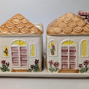 Op de afbeelding: Een keramische room- en suikerpot in de vorm van een huis op een rechthoekige schaal. De witte huizen hebben bruine pannendaken, boogvensters en bloemmotieven. De suikerpot heeft een schoorsteen.