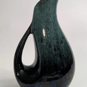 May include: A ceramic pitcher with a tall, curved spout and an integrated handle that creates an open loop. The surface features a mottled glaze transitioning from a deep, dark blue at the base to a lighter, speckled teal green toward the top rim.