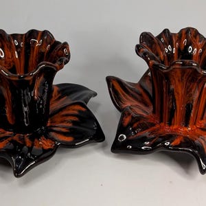 May include: A pair of ceramic candle holders shaped like blooming flowers with a glossy finish. The glaze features a marbled pattern of deep black and vibrant orange, with ruffled petal edges and a central cylindrical opening for a candle.