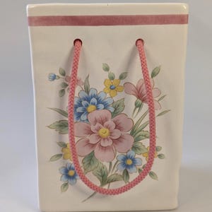 Peut inclure: Un vase rectangulaire en céramique blanche en forme de sac cadeau avec une bande horizontale rose en haut. L'avant présente une illustration florale avec des fleurs roses, bleues et jaunes et des feuilles vertes. Deux anses en corde tressée rose.
