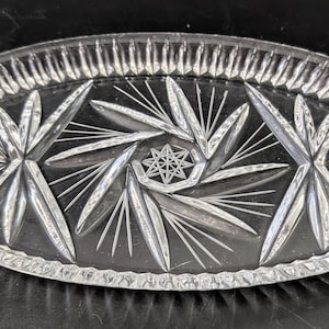 Puede incluir: Plato de vidrio transparente de forma ovalada con un diseño de estrella. El plato presenta un patrón detallado de líneas radiales y formas de estrella, con un borde texturizado. Una pieza decorativa para servir o exhibir.