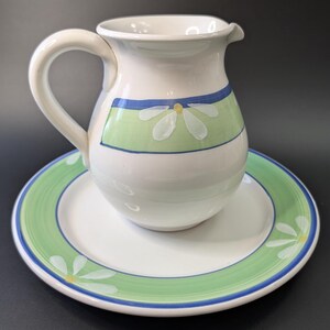May include: White ceramic jug and plate set. The jug has a curved handle and a spout. The jug and plate are decorated with a green and blue band with white daisy designs. The plate is round.