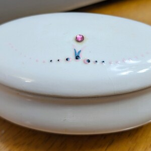 Peut inclure: Une boîte à bijoux ovale en céramique blanche avec un motif floral rose et bleu et un strass rose au centre.