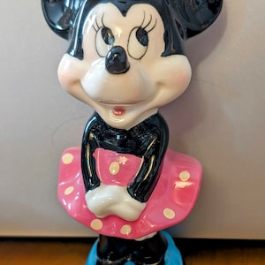 以下が含まれることがあります： ピンクのドット柄のピンクのドレス、黄色の花が付いたピンクの帽子、青い靴を履いたミニーマウスのセラミック製フィギュアです。