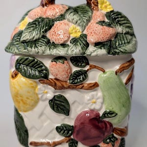 May include: A white ceramic jar with a lid, decorated with colourful, raised fruit and foliage. The lid features strawberries, green leaves, and yellow flowers. The jar's sides display a variety of fruits, including pears, apples, and lemons, with a woven basket design.