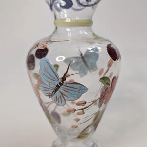 Puede incluir: Un jarrón de cristal transparente con un borde ondulado de color amarillo pálido y un cuello estrecho. El cuerpo presenta mariposas de color azul claro pintadas a mano, pequeños capullos de flores rosas, hojas verdes y detalles circulares.