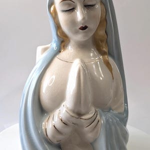 Peut inclure: Une figurine en céramique d'une femme vêtue d'une robe bleue, les mains jointes en prière. La figurine est blanche avec des accents dorés.