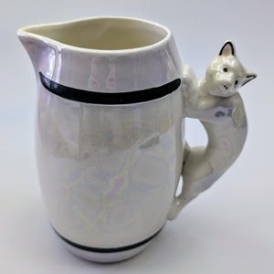 May include: White ceramic jug with a black stripe around the top and bottom. The handle is shaped like a cat with a white and black design.