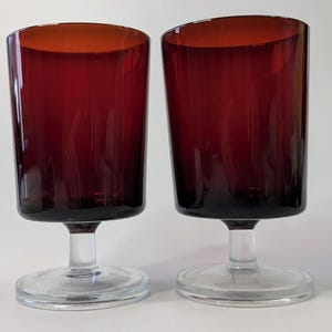 May include: Two ruby red stemmed glasses. The glasses have a clear base and stem. The cylindrical glasses have a slight taper. The glass is a deep red colour, and the base is clear.