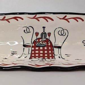 May include: A white ceramic serving platter with a black scalloped edge and a red and black cafe design. The platter features a red and white checkered tablecloth, two chairs, and the word "CAFE".