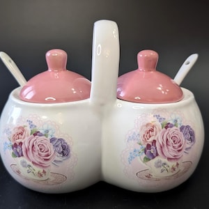 Peut inclure: Un double service à condiments en céramique blanche avec couvercles roses et une poignée. Des motifs floraux de roses et de violettes décorent les côtés. Chaque compartiment a une petite cuillère blanche.