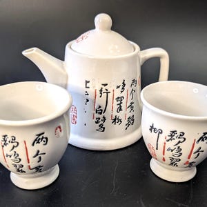 May include: A white ceramic tea set consisting of a teapot with a lid and two matching cups. Each piece features black Chinese calligraphy characters and small red stamp marks. The teapot has a curved handle and a spout, while the cups have a rounded base.