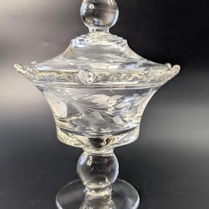 Könnte beinhalten: Klare Bonbonniere aus Glas mit Deckel. Die Schale hat einen Sockel und ein dekoratives Blumenmuster, das in das Glas geätzt ist. Der Deckel ist mit einer tropfenförmigen Spitze versehen.