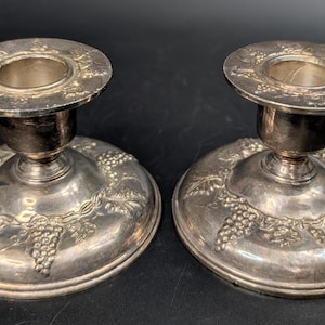 May include: A pair of silver-toned metal candle holders featuring a wide, circular base and a central socket. The surface of each holder is decorated with an embossed pattern of grape clusters and vine leaves, extending from the base to the rim.