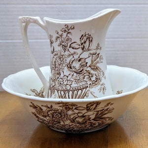 Peut inclure: Un ensemble de pichet et de bol en céramique blanche avec un motif floral brun. Le pichet a une poignée et un bec verseur. Le bol est rond et a un bord large.