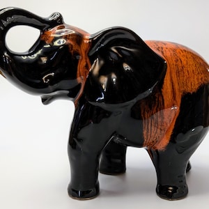 Pode incluir: Figurinha de elefante de cerâmica preta e laranja com a tromba levantada. O elefante está em pé sobre as quatro patas e tem um acabamento brilhante.