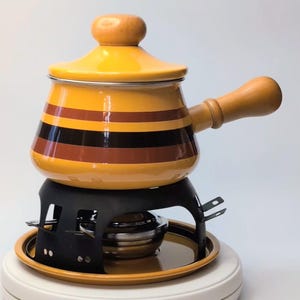 Può includere: Una pentola per fondue in smalto giallo con strisce marroni e nere e un manico in legno, appoggiata su un supporto in metallo nero con un bruciatore.