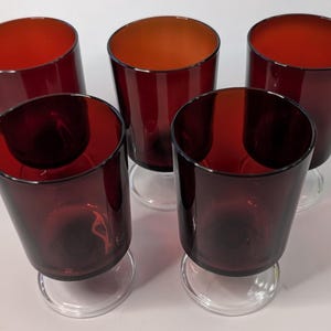 Puede incluir: Cinco copas de vino de color rojo rubí con base transparente. Las copas tienen forma cilíndrica con una ligera inclinación, y el interior de las copas tiene un tono naranja más claro. Las copas están dispuestas en círculo.