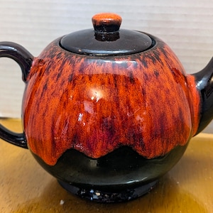 May include: A ceramic teapot with a rounded body, featuring a striking colour scheme. The top and base are glossy black, while the central portion showcases a vibrant blend of red and orange hues. The teapot includes a lid and a curved handle.